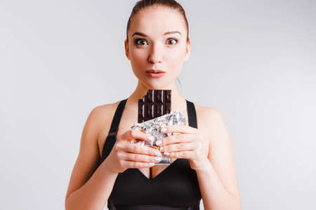 Beautiful fitness girl on a light gray background with chocolate. The concept of dealing with the...