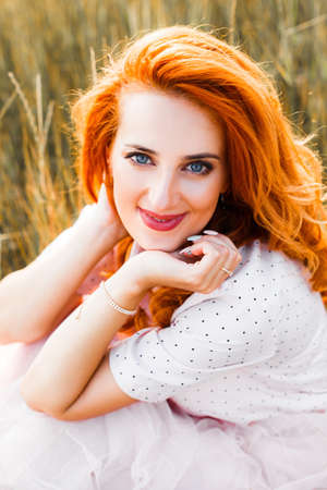 Portrait of a young red-haired woman in a tall yellow grass at sunset.