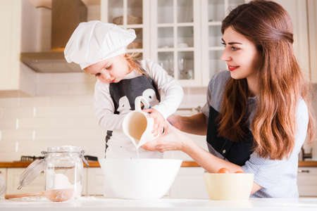 The little daughter in the chefs hat and apron and her mother prepare baking in the bright, classic...