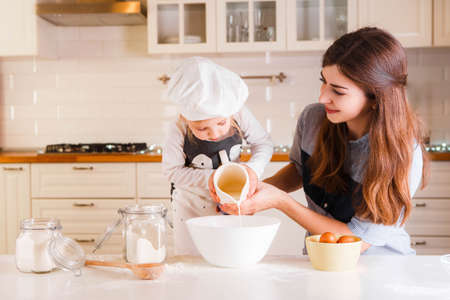 The little daughter in the chefs hat and apron and her mother prepare baking in the bright, classic...