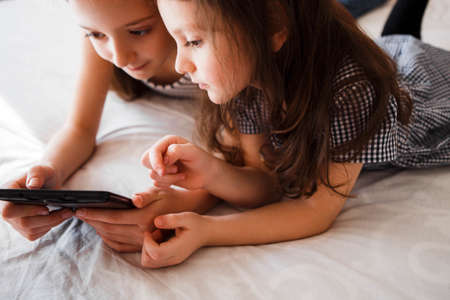 Children using digital gadgets at home. Little girls playing games on the tablet lying on the bed in...