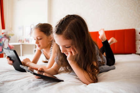 Children using digital gadgets at home. Little girls playing games on the tablet lying on the bed in the bedroom.の写真素材