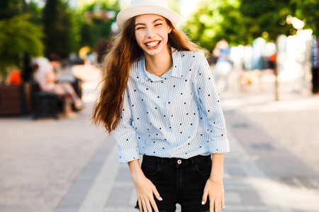Emotional girl teenager in a hat walks through the summer city. Debonair caucasian girl enjoying photoshoot on city background.の写真素材
