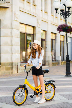 Girl teenager in a hat with a yellow bike walks through the summer city.の写真素材
