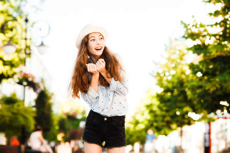Emotional girl teenager in a hat walks through the summer city. Debonair caucasian girl enjoying...