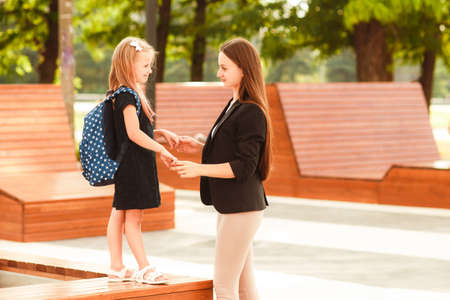 Mom leads her daughter to school. Return to school. Woman and girl with backpack behind the back. Beginning of lessons. First day of fall.の写真素材