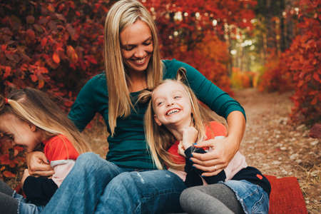 Blonde mother and her twin daughters are walking in the autumn forest among red trees.の写真素材