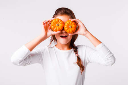 A beautiful girl on a light gray background is fooling around and grimacing with small pumpkins for a Halloween.の写真素材