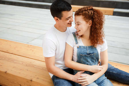 A young couple is sitting on a bench in a modern city, hugging and smiling.の写真素材