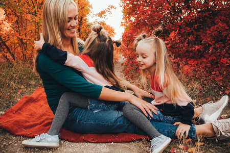 Blonde mother and her twin daughters are walking in the autumn forest among red trees.の写真素材