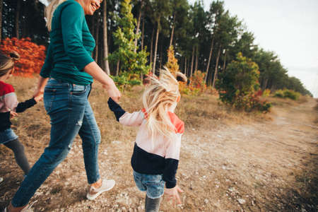 Blonde mother and her twin daughters are walking in the autumn forest among red trees.の写真素材