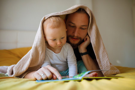 Dad with a beard and long hair reads a book to a kid. home leisure.の写真素材