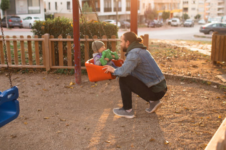 Dad and child are playing on the playground. Swing and slide rides. Sunny day.の写真素材