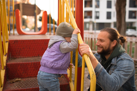 Dad and child are playing on the playground. Swing and slide rides. Sunny day.