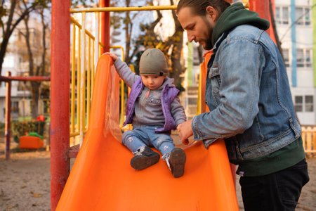 Dad and child are playing on the playground. Swing and slide rides. Sunny day.の写真素材