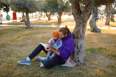 Dad and baby read a book under a tree in the park. mediterranean landscape.の写真素材