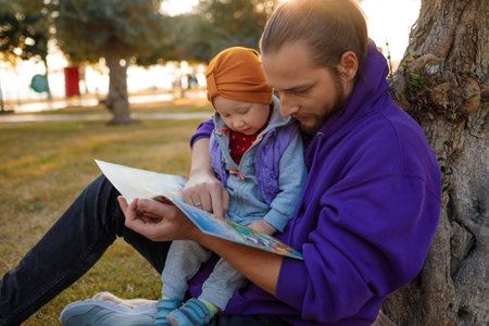 Dad and baby read a book under a tree in the park. mediterranean landscape.の写真素材