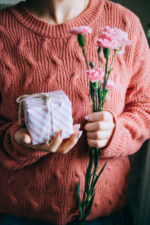 Woman in coral woolen sweater girl holding gift box with ribbon and fresh green leaves and blooming pink flowersの写真素材