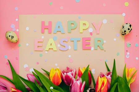 Easter concept. Happy easter paper cut inscription with pink and red tulips and quail eggs on pink background. Copy space, flat lay.の写真素材