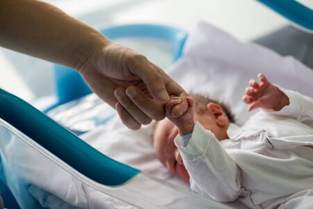 Beautiful newborn baby boy, laying in a small crib holding his father finger in prenatal hospitalの写真素材
