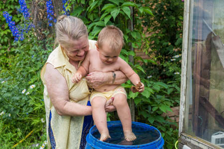 Grandmother rinses the feet of the baby in a barrel of water in the gardenの写真素材