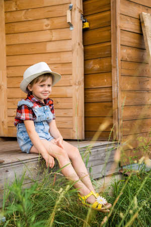 Adorable Children girl, kid cowgirl sitting on the porch of a wooden houseの写真素材