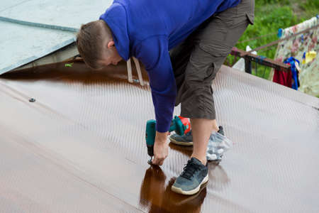 A man holds a screwdriver in his hands while standing on the roof of a village house. Securing the new polycarbonate roof canopy with thermal washersの写真素材