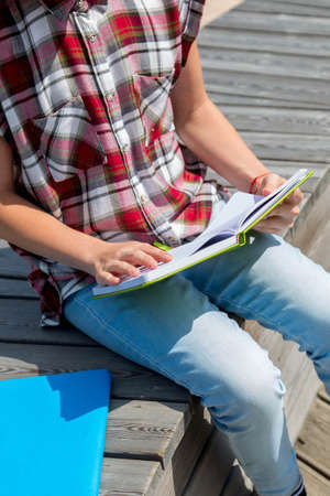 Lifestyle image of pretty school girl sitting on the bench in public park and making notes to her...