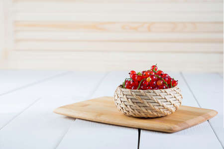 Ripe tasty bright red currants in a wicker basket on a cutting board on a white wood background