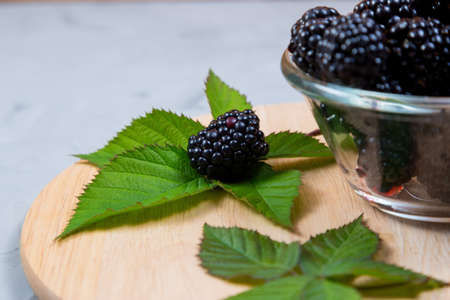 ripe blackberries with leaves in a glass bowl on a bamboo cutting board on a concrete background,...