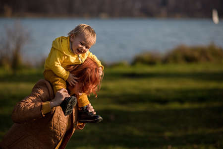 .Baby todler in a yellow jacket laughs while sitting on the neck of the mother against of the autumn...