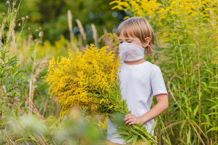 .A little girl in a medical mask holds a huge bouquet of yellow autumn flowers in a field. Coronavirus prevention, keep social distance and keep wearing masksの写真素材