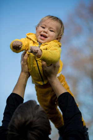 .Dad throws a baby in a yellow jacket into the air, the kid laughs and has fun, an autumn park, an Indian summer.の写真素材