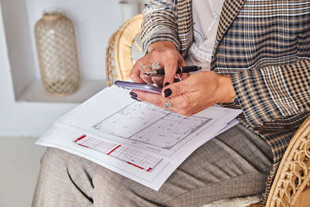 Woman interior designer with interior plans for a new project holds a mobile phone in her hands,...