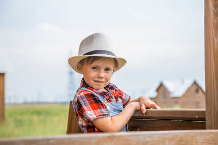 Cute little girl in a cowboy hat, dressed in a denim shorts and red white shirt, spends time in the village in summer, stands near a wooden house.の写真素材