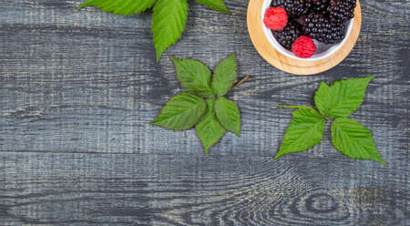 ripe blackberry with leaves on a wooden cutting board in a white ceramic bowl on dark blue wooden...