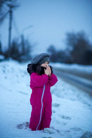 Little girl warms her hands with her breath on a winter evening near the roadの写真素材