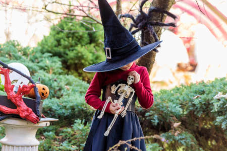 Little girl in a witch costume holding a skeleton on a halloween party in the gardenの写真素材