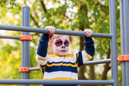 A little preschool girl with Painted Face, looking at the camera on the playground, celebrates Halloween or Mexican Day of the Dead ..の写真素材