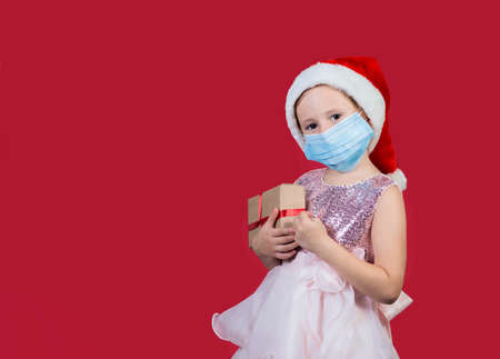 Adorable caucasian little girl wearing medical mask in santa hat with gift box celebrating happy New Year isolated on red background. Merry Christmas presents shopping sale.の写真素材