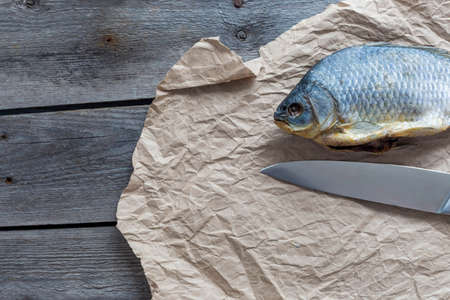 Salted Dry fish vobla with a sharp knife on crumpled craft paper on wooden background, delicious beer snack, close-up.の写真素材