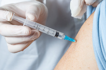 A doctor holds a syringe with a vaccine in his hands and injects the vaccine into the shoulder of an elderly patient in a clinic.の写真素材