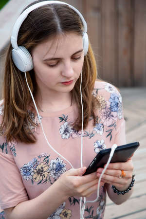 A young girl 20 years old Caucasian appearance looks into her mobile phone, writes a text message while sitting on a wooden podium in the park on a summer day.の写真素材