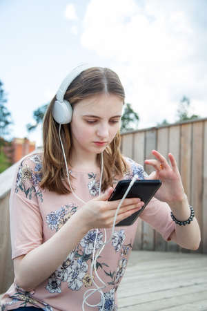 A young girl 20 years old Caucasian appearance looks into her mobile phone, writes a text message while sitting on a wooden podium in the park on a summer day.の写真素材