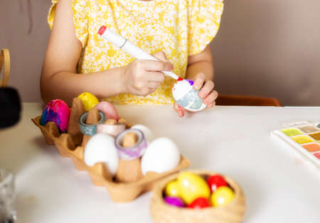 A close-up on the hands of a small Caucasian girl 5 years old paints eggs with special markers paints for the Christian spring holiday of Easter. Girl dressed in a yellow floral dressの写真素材