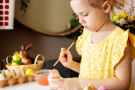 A small Caucasian girl of 5 years old paints eggs with special water paints for the Christian spring holiday of Easter. The girl is dressed in a yellow floral dress and has ponytails.の写真素材
