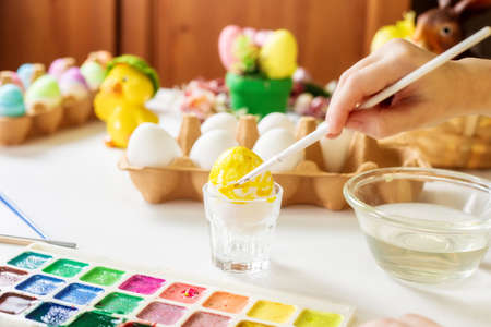A close-up on the hands of a small Caucasian girl 5 years old paints eggs with special water paints for the Christian spring holiday of Easter. Girl dressed in a yellow floral dressの写真素材