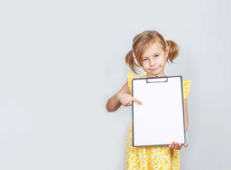 Little Caucasian girl holding a clipboard with a blank paper with copy space for inscription, advertisement posing against studio wall. Advertising conceptの写真素材