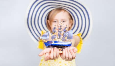 A Little Caucasian girl in a wide-brimmed hat holds out a toy boat forward. Focus on the foreground. The concept of summer vacation, travel, opening of the beach season ..の写真素材