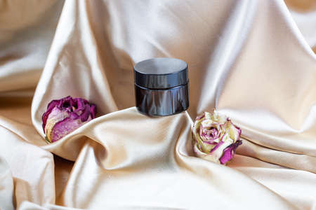 A dark glass makeup container lies on folds of silk satin, surrounded by dried rose flowersの写真素材